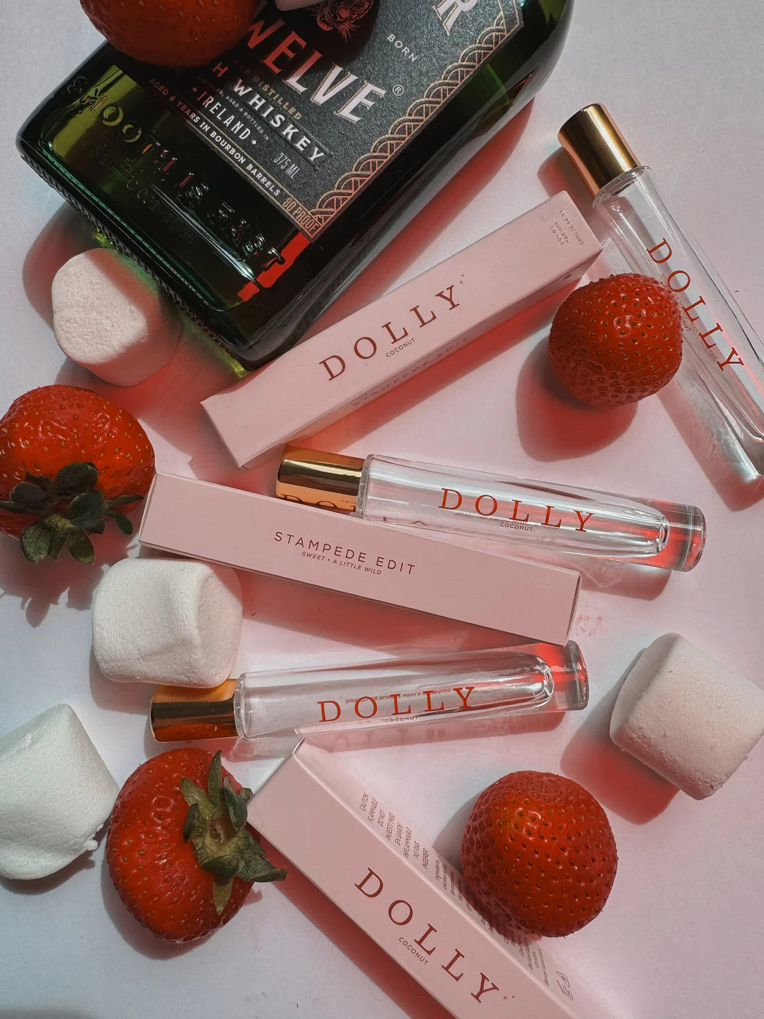 A styled flat-lay of Dolly perfume travel atomizers and their pink boxes, arranged with strawberries and marshmallows on a light pink background. A bottle of Jim Beam whiskey is partially visible in the top left as a prop.