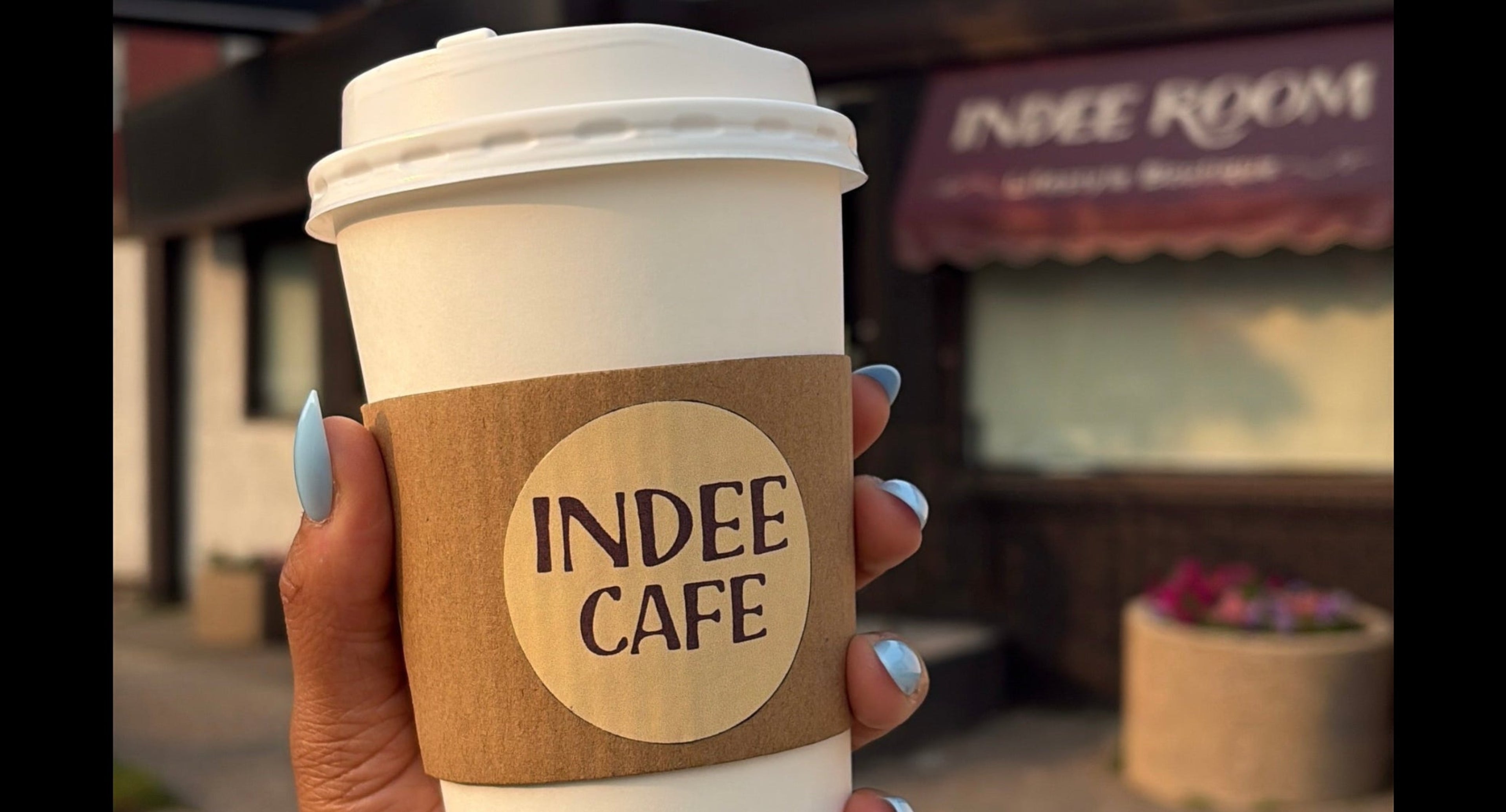 A hand holding a white paper coffee cup with a brown paper sleeve printed with 'INDEE CAFE' in black text, with a blurred storefront in the background.