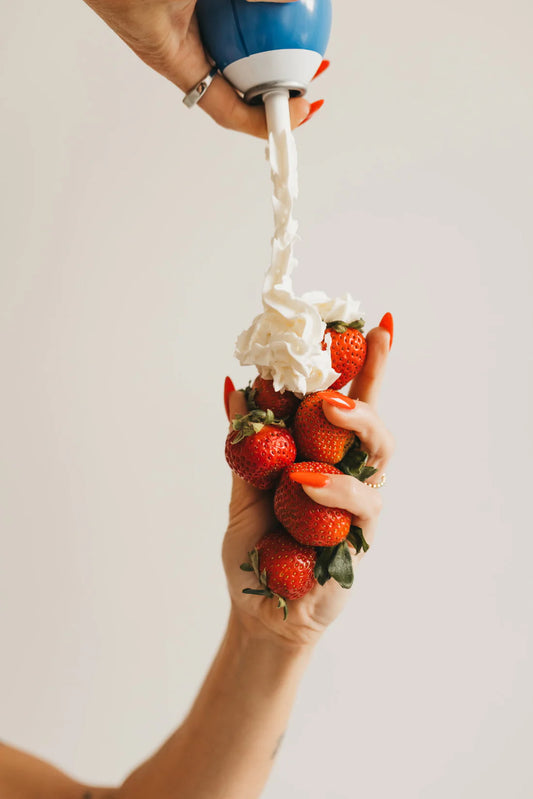A close-up of a hand holding a cluster of fresh red strawberries while another hand dispenses whipped cream from a blue and white canister onto the strawberries against a plain light background.